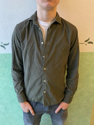 Mörkgrön Manchester skjorta från Riley - Stilren mörkgrön skjorta från Riley med klassisk krage och vita knappar framtill. Skjortan är långärmad och har en avslappnad passform. Tillverkad i mjuk bomull som känns skön mot huden. Perfekt att bära över en t-shirt för en casual look.