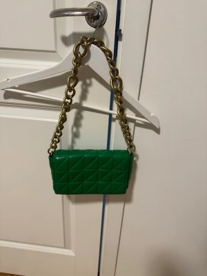 Grön quiltad kuvertväska med kedja - Snygg grön kuvertväska i quiltat konstläder med en chunky guldfärgad kedja som axelrem. Väskan har en rektangulär form och coolt rutmönster på både fram- och baksida. Perfekt för dig som vill sticka ut med en färgklick till din outfit.