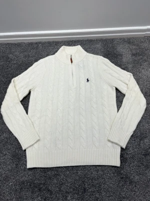 Vit kabelstickad tröja Polo Ralph Lauren - Kabelstickad vit tröja från Polo Ralph Lauren med halv dragkedja och hög krage. Tröjan har ribbade muddar och nederkant samt den klassiska broderade loggan på bröstet. Perfekt för dig som gillar stilrena och tidlösa plagg.