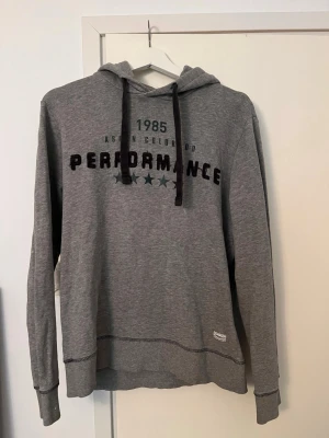 Grå vintage hoodie  - Snygg grå hoodie med svart tryck 'Performance 1985' och stjärndetaljer på bröstet. Hoodien har huva med svarta snören och ribbade muddar vid ärmslut och nederkant. Perfekt för en avslappnad och sportig stil. 