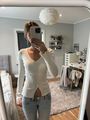 Vit ribbad långärmad tröja - Säljer en vit ribbad långärmad tröja med djup v-ringning och cool detalj med öppning framtill. Tröjan är figurnära och har en modern, stilren look som passar perfekt till jeans. Materialet ser ut att vara mjukt och stretchigt.
