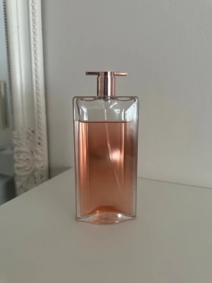 Lancôme Idôle Eau de Parfum - Säljer en Lancôme Idôle Eau de Parfum i en elegant, rektangulär glasflaska med roséguldigt lock och ljusrosa doftvätska. Flaskan har en modern och tunn design som känns lyxig och stilren – perfekt för dig som gillar exklusiva parfymer.