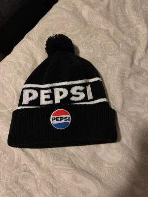 Åre mössa - Säljer en svart stickad mössa från Pepsi med vit logga och text framtill. Mössan har en klassisk tofs på toppen och en bred uppvikt kant med Pepsi-märke i rött, vitt och blått. Perfekt för dig som gillar retrostil och vill sticka ut lite extra.