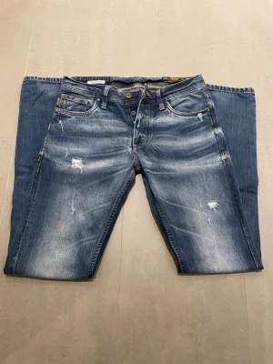 Jack and Jones jeans - Säljer ett par blå jeans med slitningar och snygg tvätt. Jeansen har knäppning med flera knappar framtill, fem fickor och kontrastsömmar. Bakfickorna har dekorativa sömmar och det finns en läderpatch i midjan. Perfekta för en avslappnad stil. Jeansens storlek står inte i men jag skulle säga att dom är 30/32. Dom har ett väldigt bra pris men går att diskutera. Skriv vid minsta fundering!!🤩🤩