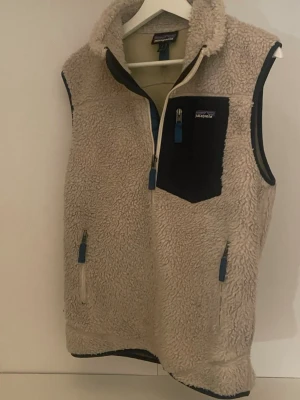 Patagonia fleeceväst  - Säljer min supermysiga fleeceväst från Patagonia i storlek S. Klassisk teddy-/pilefleece i beige med mörkblå detaljer och bröstficka med logga. Perfekt som lager på lager under höst/vinter eller som ytterplagg svala dagar