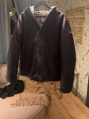 Lila vindjacka från Colmar - Snygg mörkblå dunvindjacka från Colmar med huva och dragkedja framtill. Jackan har två fickor med dragkedja och en diskret logga på ärmen. Perfekt för blåsiga dagar och enkel att matcha med olika outfits.