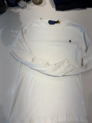 Vit tröja från Polo Ralph Lauren - Vit långärmad tröja från Polo Ralph Lauren med diskret blå logga broderad på bröstet. Tröjan har rund halsringning och är tillverkad i ett ribbat, mjukt material som ger en avslappnad och stilren look.
