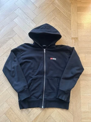 Svart hoodie från Diesel  - Svart hoodie från Diesel med dragkedja framtill och stor logga på ryggen samt mindre logga på bröstet. Klassisk modell med huva och känguruficka. Tillverkad i mjukt bomullsmaterial och har ribbade muddar vid ärmslut och nederkant. Perfekt för en avslappnad streetwear-look.