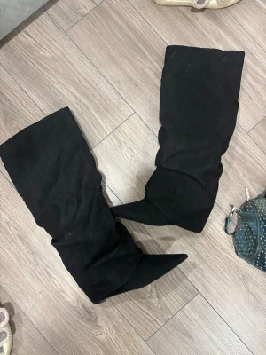 Svarta slouchy stövlar med spetsig tå - Säljer ett par höga svarta stövlar med slouchy passform och spetsig tå. Stövlarna har en bredare skaft och är tillverkade i ett mjukt mockaliknande material. Perfekta för att lyfta vilken outfit som helst med sin edgy look. 9cm