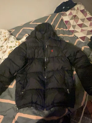 Svart pufferjacka från Polo Ralph Lauren - Säljer en svart pufferjacka från Polo Ralph Lauren med klassisk röd logga på bröstet. Jackan har dragkedja framtill, två sidofickor med dragkedja och en bröstficka med dragkedja. Fylld för extra värme och har hög krage för att skydda mot kyla. Pris kan diskuteras .