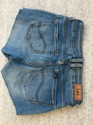 Blå jeansshorts från Lee - Säljer ett par klassiska blå jeansshorts från Lee med råa kanter och fem fickor. Shortsen har slitna detaljer och Lee-logga på bakfickan. Perfekta för sommaren och har en avslappnad passform. Shortsen har några tecken på andvändning som man kan se på bild 5🩵