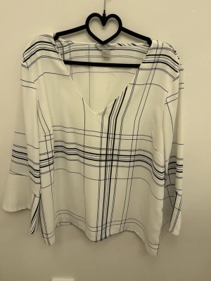 Vit blus med svart rutmönster H&M - Snygg vit blus från H&M med svart rutigt mönster och V-ringning. Blusen har trekvartsärm och är gjord i ett lätt, mjukt material som känns skönt mot huden. Perfekt för en trendig och stilren look.