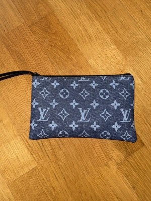 Louis Vuitton monogram kuvertväska - Snygg kuvertväska från Louis Vuitton i blått denimliknande tyg med klassiskt monogrammönster i vitt. Väskan har en smidig dragkedja upptill och en svart handledsrem. Perfekt för att bära det viktigaste med stil.