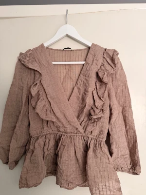 Beige volangblus från ONLY - Supersöt beige blus från ONLY med volangdetaljer och omlott-skärning. Blusen har långa ärmar och resår i midjan.