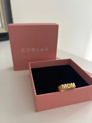 Guld ring MOM  Edblad - Snygg guldfärgad ring från Edblad med texten MOM i stora bokstäver framtill. Ringen har en kantig, modern design och är tillverkad i metall med en blank finish. Perfekt present inför mors dag eller någon speciell. Kan även vändas så det blir WOW. 