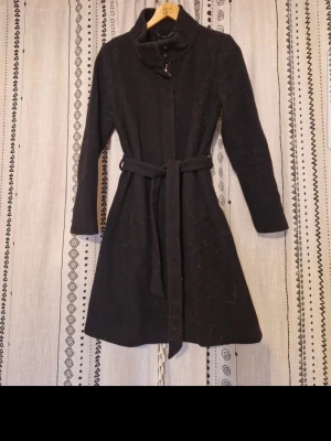 Svart kappa med knytskärp - Stilren svart kappa med hög krage och knytskärp i midjan. Kappan har en figurnära passform, långa ärmar och slät yta. Perfekt för kyligare dagar och enkel att matcha med olika outfits.