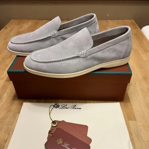 Ljusgrå loafers från Loro Piana - Säljer ett par ljusgrå loafers från Loro Piana i mjukt mockamaterial. Skorna har en klassisk och stilren design med vita sömmar och ljus sula. Perfekta för dig som gillar en clean och elegant look. Originalkartong och dustbag medföljer.