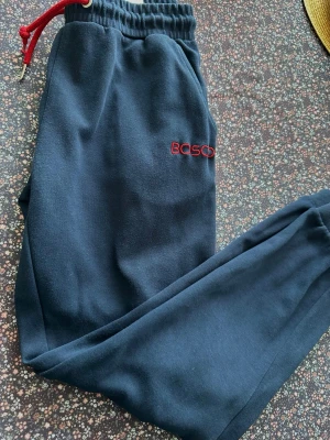 Mörkblå mjukisbyxor från Bosco M - Mörkblå mjukisbyxor från Bosco med röd dragsko i midjan och broderad logga på vänster ben. Byxorna har resår i midjan och är tillverkade i mjukt, bekvämt material. Perfekta för chill eller träning.