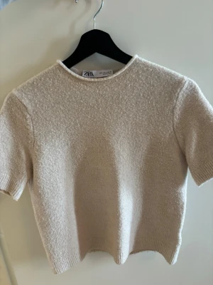 Beige stickad topp från Zara - Mysig beige stickad topp från Zara med korta ärmar och rund hals. Toppen har en enkel, stilren design och är gjord i ett mjukt material som känns skönt mot huden. Perfekt att matcha med jeans eller kjol för en clean look.