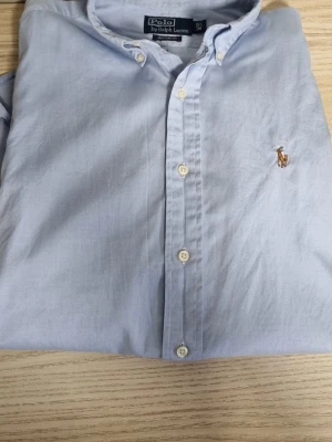 Ljusblå skjorta från Polo Ralph Lauren - Klassisk ljusblå skjorta från Polo Ralph Lauren med broderad logga på bröstet. Skjortan har knappar framtill och lång ärm. Tillverkad i mjuk bomull för en bekväm känsla och stilren look.