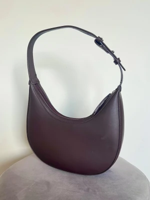 Vintage style brun shoulder bag baguette bag 90s/Y2K aesthetic (ny) - Snygg mörkbrun/chokladbrun axelväska i  halvmåneformad design. Minimalistisk, 90-tal och trendig. Aldrig använd. Vet ej var den kommer ifrån.  Höjd: ca 15-16cm Band: ca 44cm