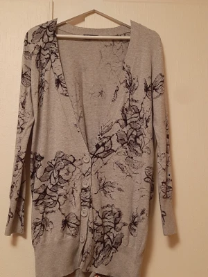 Grå kofta med blommigt mönster - Snygg grå kofta med svart blommigt mönster och stora knappar framtill. Långärmad och i mjukt, tunt material som sitter skönt på kroppen. Perfekt att slänga över en topp för en chill och stilren look.