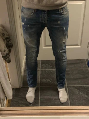 Blå jeans från Taylor Tweed - Säljer ett par blå jeans från Taylor Tweed med slitningar och distressed detaljer på både fram- och baksida. Jeansen har raka ben, knappgylf och klassisk femficksmodell. Materialet är denim i bomull och passformen är smal. Priset kan diskuteras vid snabbt köp. Dessa är knappast använda så de är näst intill nya och är slim