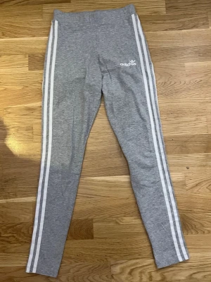 Grå leggings från Adidas - Grå leggings från Adidas med vita ränder längs benen och Adidas-logga på höften. Tight passform och hög midja. Materialet är mjukt och stretchigt. 