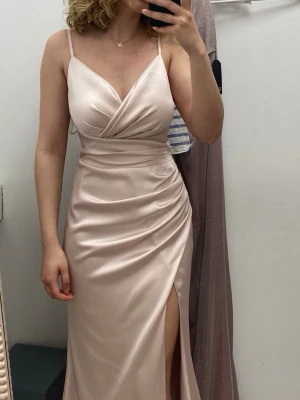 Rosa långklänning med slits - Elegant långklänning i Rosa satinliknande material med smala axelband, omlottliv och hög slits framtill. Klänningen har en figurnära passform och v-ringad urringning som ger en stilren och lyxig känsla. Använd 1 gång.