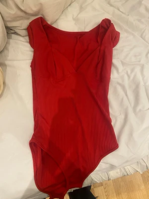 Röd ribbad body med v-ringning - Säljer en snygg röd body med ribbad struktur och djup v-ringning. Bodyn har korta ärmar och markerade kupor framtill för en smickrande passform. Perfekt att styla till jeans eller kjol för en trendig look.