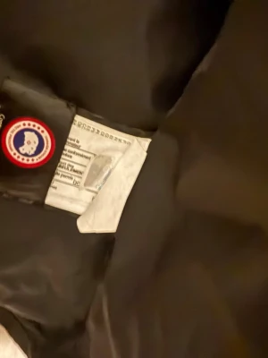 Svart dunjacka från Canada Goose - Säljer en svart dunjacka från Canada Goose med klassisk patch på insidan. Jackan har dragkedja och tryckknappar, samt en stilren design som passar till det mesta. Materialet är slitstarkt och fyllningen består av dun för maximal värme.