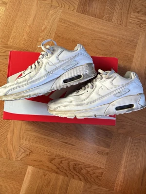 Nike Air Max 90 vita sneakers - Nike Air Max 90 sneakers i helvitt läder med klassisk synlig Air-enhet i sulan. Snygg retrodesign med meshdetaljer och tydliga Nike-loggor på sidan och hälen. Perfekta för dig som gillar streetwear och tidlös stil.