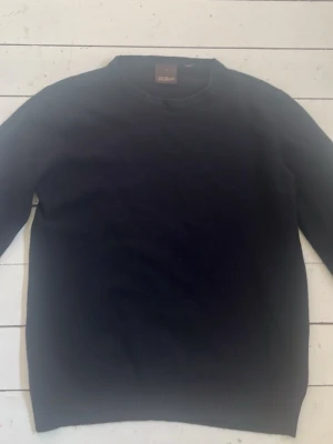 Mörkblå stickad tröja Oscar Jacobson - Klassisk mörkblå stickad tröja från Oscar Jacobson med rund halsringning och ribbade muddar vid ärmslut och nederkant. Perfekt basplagg i garderoben och enkel att matcha med olika stilar. Materialet känns mjukt och bekvämt.