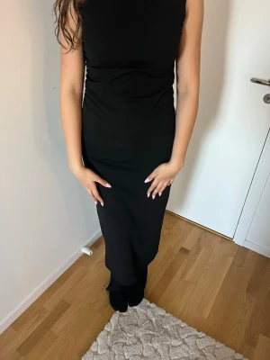 Svart ärmlös långklänning - Stilren svart långklänning med ärmlös design och rund halsringning. Klänningen har en smickrande passform och enkel siluett som ger ett elegant intryck. Perfekt för dig som gillar minimalistisk stil. Materialet känns mjukt och följsamt.