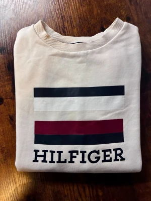 Vit sweatshirt från Tommy Hilfiger - Snygg vit sweatshirt från Tommy Hilfiger med klassiskt tryck i svart, vitt och vinrött på bröstet samt Hilfiger-logga. Tröjan har rund halsringning, ribbade muddar och är tillverkad i mjuk bomull. Perfekt för en clean och stilren look.