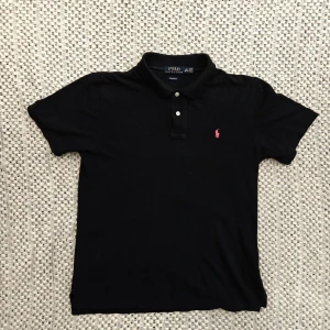 Svart pikétröja från Polo Ralph Lauren - Svart pikétröja från Polo Ralph Lauren med klassisk krage, två vita knappar och röd broderad logga på bröstet. Tillverkad i mjuk bomull och har korta ärmar för en avslappnad men stilren look. Har en väldigt fadad vita fläck vid kragen som lätt går bort i tvätt