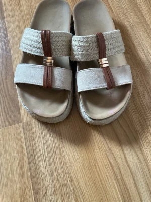 Beige slip-in tofflor med flätad detalj - Säljer ett par beige slip-in tofflor med två breda band över foten, varav det ena är flätat och det andra i tyg. Tofflorna har bruna dekorativa remmar med guldfärgade detaljer. Sulan är något upphöjd och har en naturlig look.