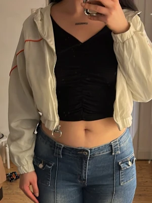 Vit cropped vindjacka med orange detaljer - Säljer en vit cropped vindjacka med dragkedja och orange detaljer längs ärmarna. Jackan har resår i midjan och muddar vid ärmsluten. Märke 2L syns på ärmen. Perfekt för en sportig och avslappnad stil.