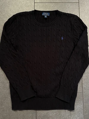 Svart kabelstickad tröja Polo Ralph Lauren - Kabelstickad svart tröja från Polo Ralph Lauren med rund halsringning och lila broderad logga på bröstet. Tröjan har långa ärmar och ribbade muddar vid ärmslut och nederkant. Perfekt för dig som gillar klassisk och stilren design. Barnstorlek XL / Herr S (se bilder för mått)