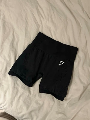 Svarta träningstights shorts Gymshark - Säljer ett par svarta högmidjade träningstights shorts från Gymshark. De har en tight passform, bred resår i midjan och ventilerande meshdetaljer längs sidorna. Perfekta för gymmet eller löpning.