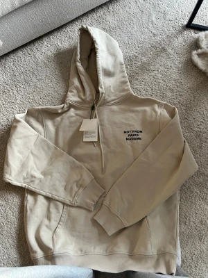 Beige hoodie Not From Paris Madame - Snygg beige hoodie från Not From Paris Madame med svart text på bröstet. Hoodien har klassisk passform, ribbade muddar och en stor huva. Materialet är mjukt och bekvämt, perfekt för en chill look. Dragkedja i halsen och diskret design gör den enkel att matcha.