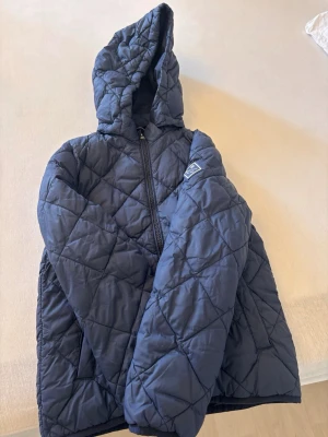 Mörkblå pufferjacka från Gant - Snygg mörkblå pufferjacka från Gant med quiltat mönster och huva. Jackan har dragkedja framtill, två fickor och en patch med Gant-logga på ärmen. Materialet är vadderat och håller värmen bra.