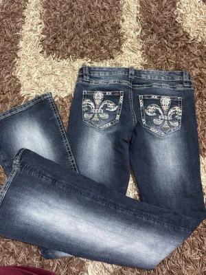 Mörkblå jeans med broderade bakfickor - Snygga mörkblå jeans med slitningar och vita broderade detaljer på bakfickorna i form av ett stort mönster. Jeansen har raka ben och kontrastsömmar. Perfekta för dig som vill sticka ut lite extra med coola detaljer.  (Tall)