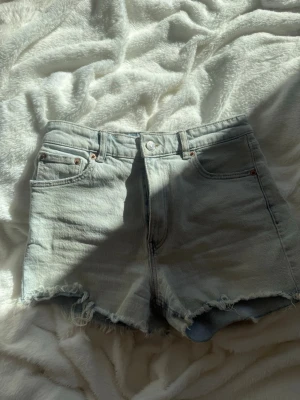 Ljusblå denimshorts med fransig kant - Säljer ett par ljusblå denimshorts med fransig rå kant och klassisk femficksdesign. Shortsen har medelhög midja och stängs med knapp och dragkedja. Perfekta för varma dagar och ger en avslappnad vibe.
