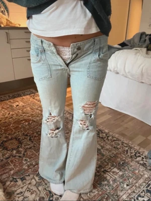 Ljusblå slitna bootcut jeans - Säljer ett par ljusblå bootcut jeans med slitningar och hål på båda benen. Jeansen har låg midja och klassiska fickor fram. Perfekta för en avslappnad och trendig look.