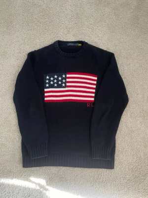 Mörkblå stickad tröja Polo Ralph Lauren - Mörkblå stickad tröja från Polo Ralph Lauren med amerikansk flagga i rött, vitt och blått på bröstet. RL-broderi i rött under flaggan. Tröjan har rund hals och ribbstickade muddar vid ärmslut och nederkant. Perfekt för dig som gillar klassisk och cool stil.