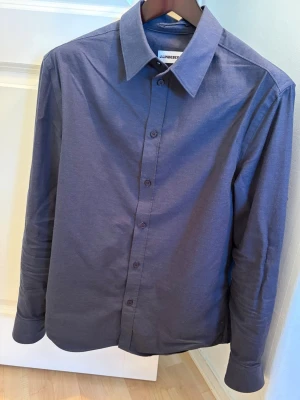 Mörkblå skjorta från J.Lindeberg - Stilren mörkblå skjorta från J.Lindeberg med klassisk krage och knäppning framtill. Skjortan har långa ärmar och är tillverkad i ett mjukt flannel material som ger en skön vibe. Perfekt för någon som gillar stillfull och modern stil.