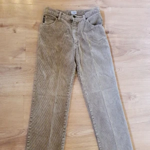 Beiga manchesterbyxor från Armani Jeans - Snygga beiga manchesterbyxor från Armani Jeans med klassisk rak passform och fem fickor. Byxorna har breda manchesterränder och detaljer som logopatch bak. Perfekta för dig som vill ha en stilren och trendig look. Storlek ca 160 cm (ca juniorstl L). Bra skick överlag dock finns det en liten lagning på knät.