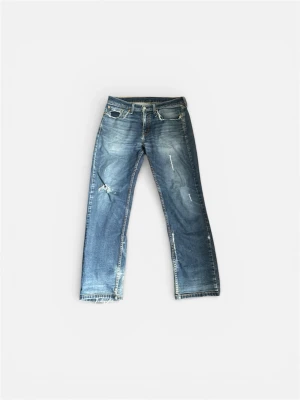 Blå slitna Levi's jeans med hål - Säljer ett par blå Levi's jeans med slitningar och hål på vänster knä och ficka. Jeansen har klassisk femficksmodell, raka ben och ljus tvätt med snyggt sliten look. Perfekta för en avslappnad och trendig stil.