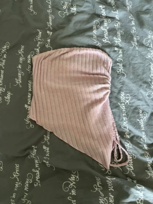 Ljusrosa ribbad bandeau tubtopp - Trendig ljusrosa bandeau tubtopp med ribbad struktur och snörning på sidan. Toppen har en asymmetrisk, spetsig nederkant och är perfekt för sommaren. Materialet känns mjukt och stretchigt, vilket gör den bekväm att bära.